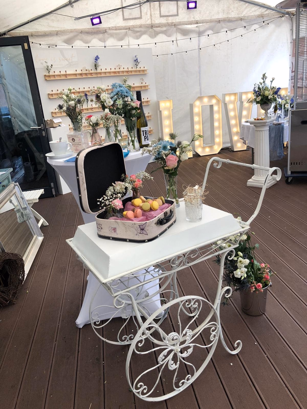 Weißer Rollwagen mit Macarons, floralen Vasen und LOVE-Leuchtbuchstaben als Empfangselement.