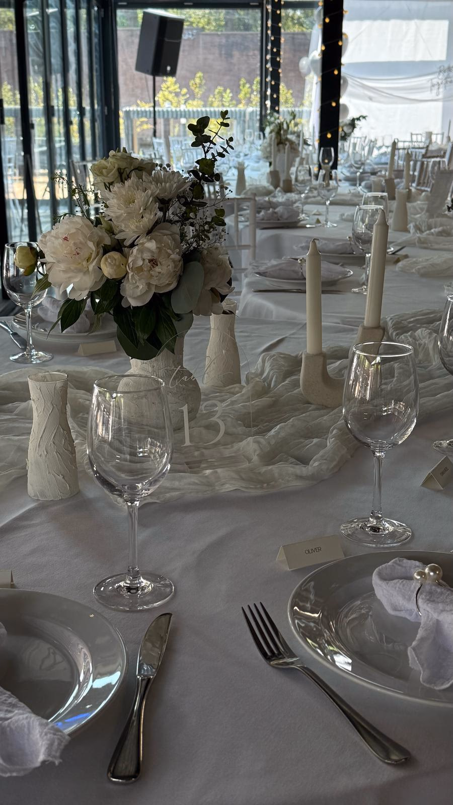 Gedeckte Rundtafel mit weißen Pfingstrosen in strukturierter Keramikvase, Stabkerzen und Cheesecloth-Läufer.